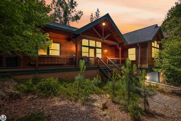 Modern wood cabin with illuminated windows and wraparound deck at dusk, surrounded by evergreen trees and landscaping.