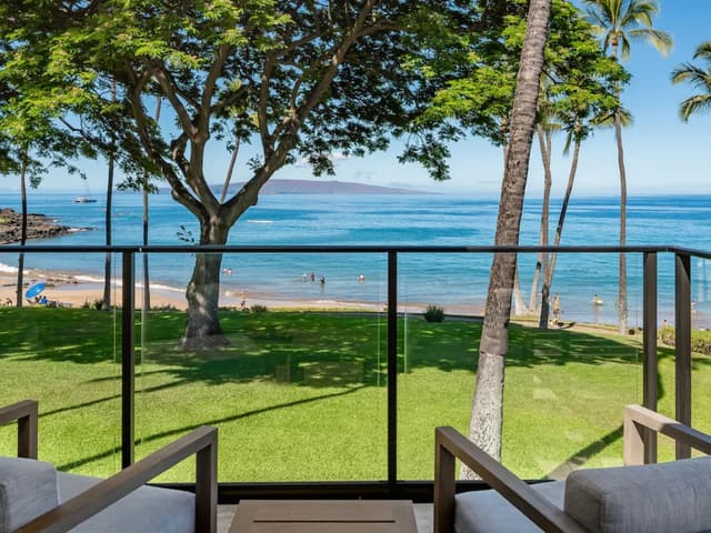 Oceanfront lanai with glass railing overlooking sandy beach, palm trees, and blue ocean with swimmers